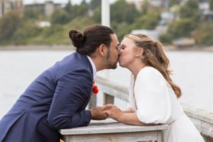 A couple shares a romantic kiss while leaning over a stone wall near the waterfront on their wedding day.