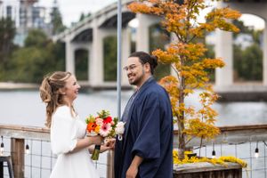 A couple smiles at each other on a dock during autumn with vibrant orange leaves and a bridge in the background.