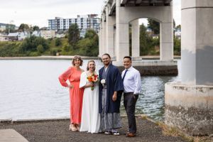 Small wedding party poses beneath a bridge near waterfront with city buildings visible in background.