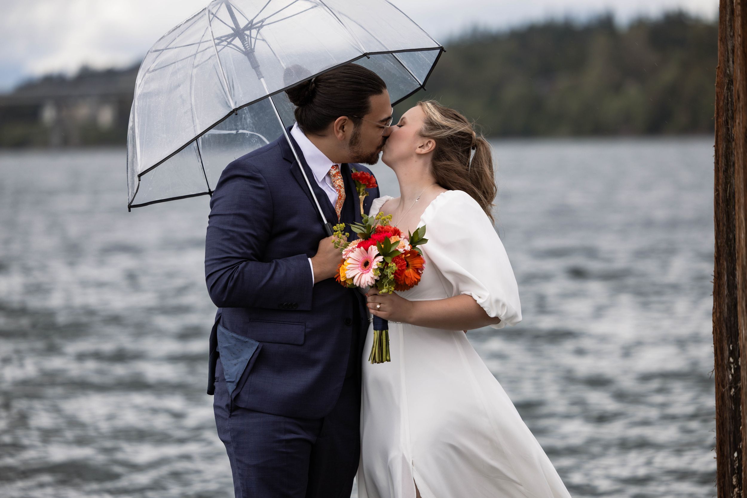 A couple shares a romantic kiss under a clear umbrella by a lake, with the bride holding a colorful bouquet.