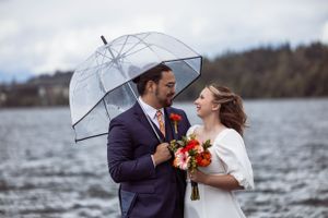 A couple shares a romantic moment under a clear umbrella by a lake, with the bride holding coral-colored flowers.
