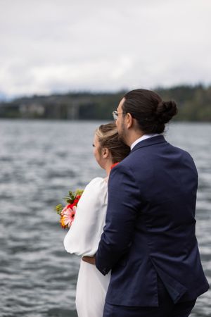 A couple in wedding attire stand by a lake, with the bride in white holding flowers and groom in navy suit.