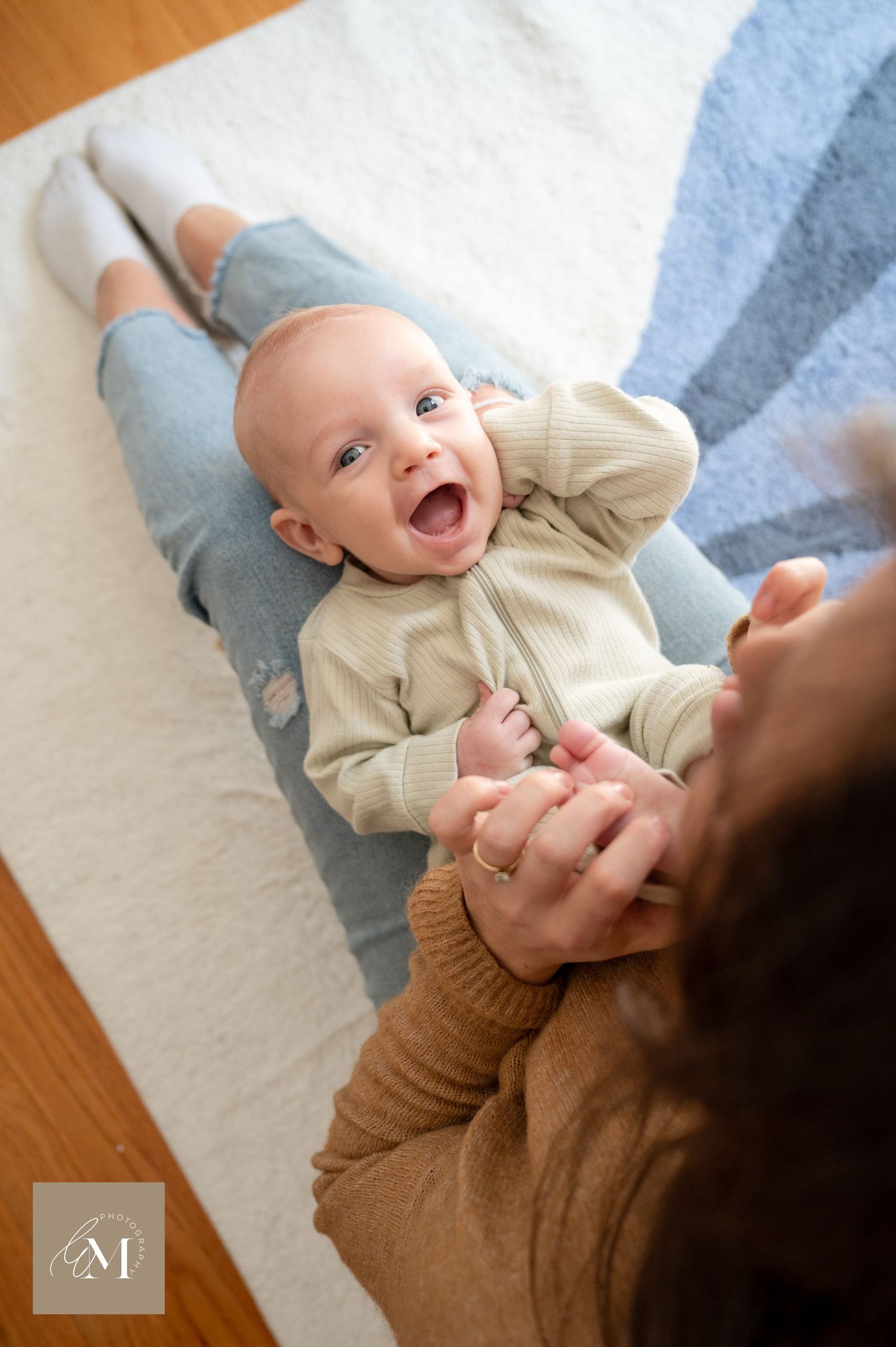 Happy baby wearing cream sweater lying on floor blanket while being tickled with laughter.