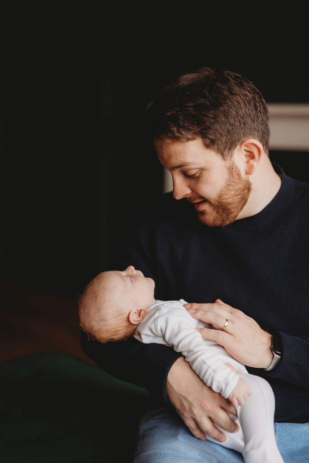 A tender moment between a parent in a navy sweater gazing adoringly at their sleeping infant in white clothes.