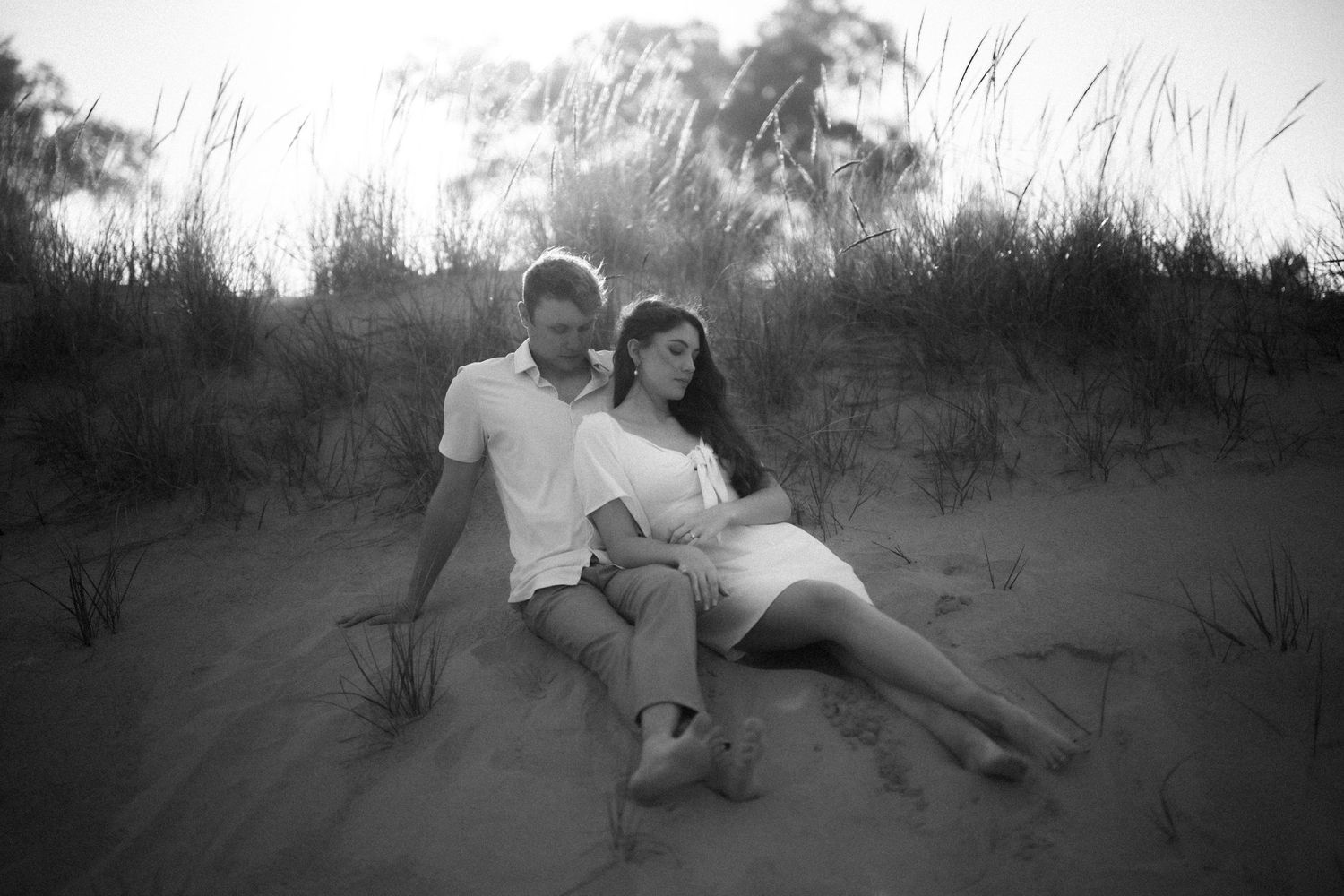 A dreamy black and white photograph of a couple lounging together on sand dunes at dusk.