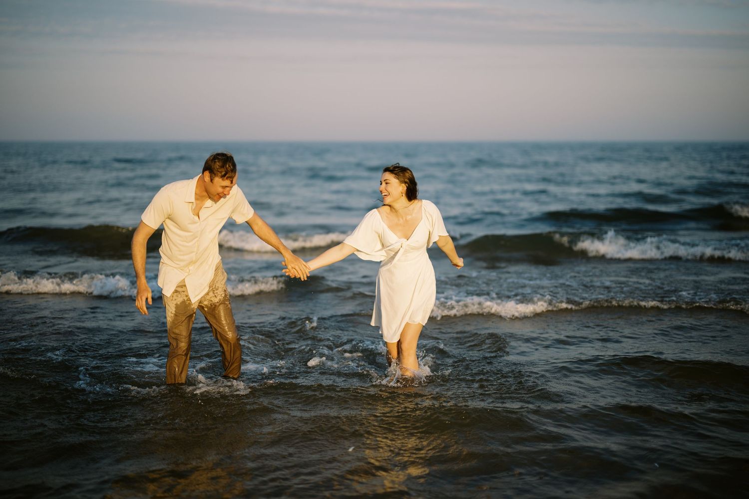 A couple in white outfits holding hands while wading in ocean waves at sunset.