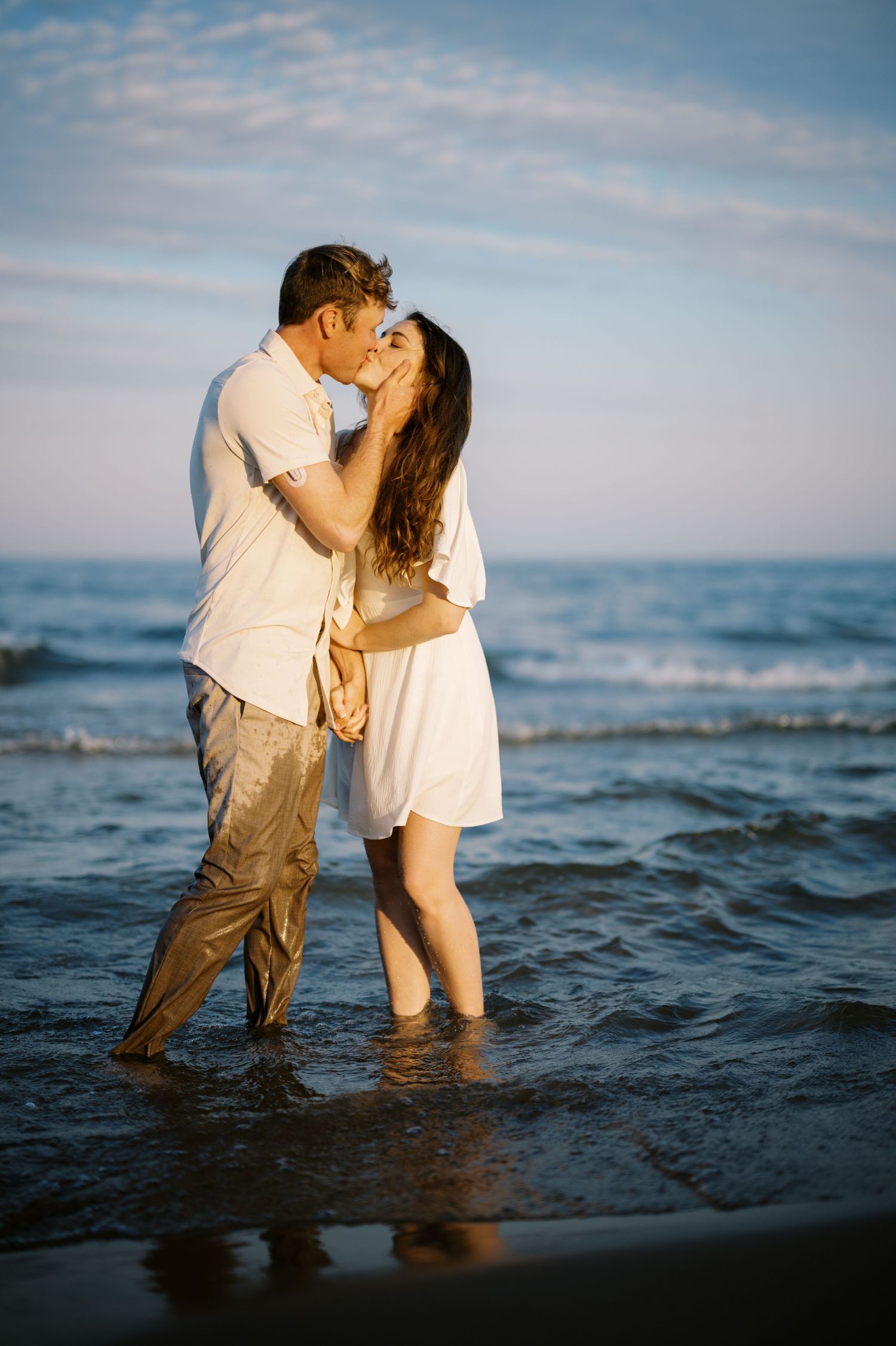 A couple shares a romantic kiss while standing in shallow ocean water at sunset.
