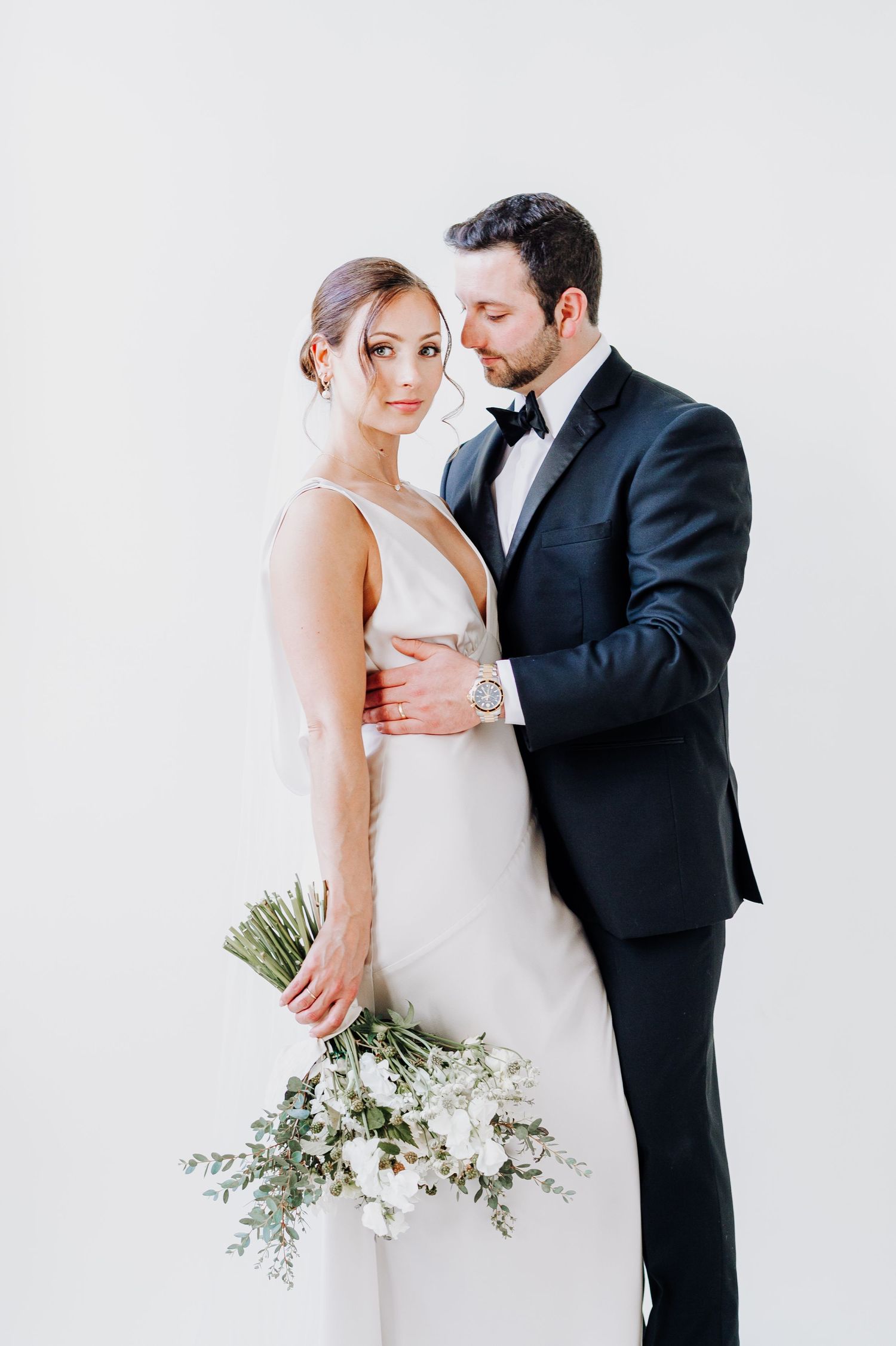Elegant wedding couple poses together against white background with bride holding delicate white and green bouquet.