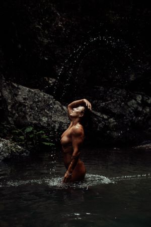 A figure creates an artistic water arc while standing in a dark forest pool.
