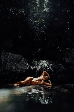 Artistic nude photography in a secluded forest pool with dramatic lighting.