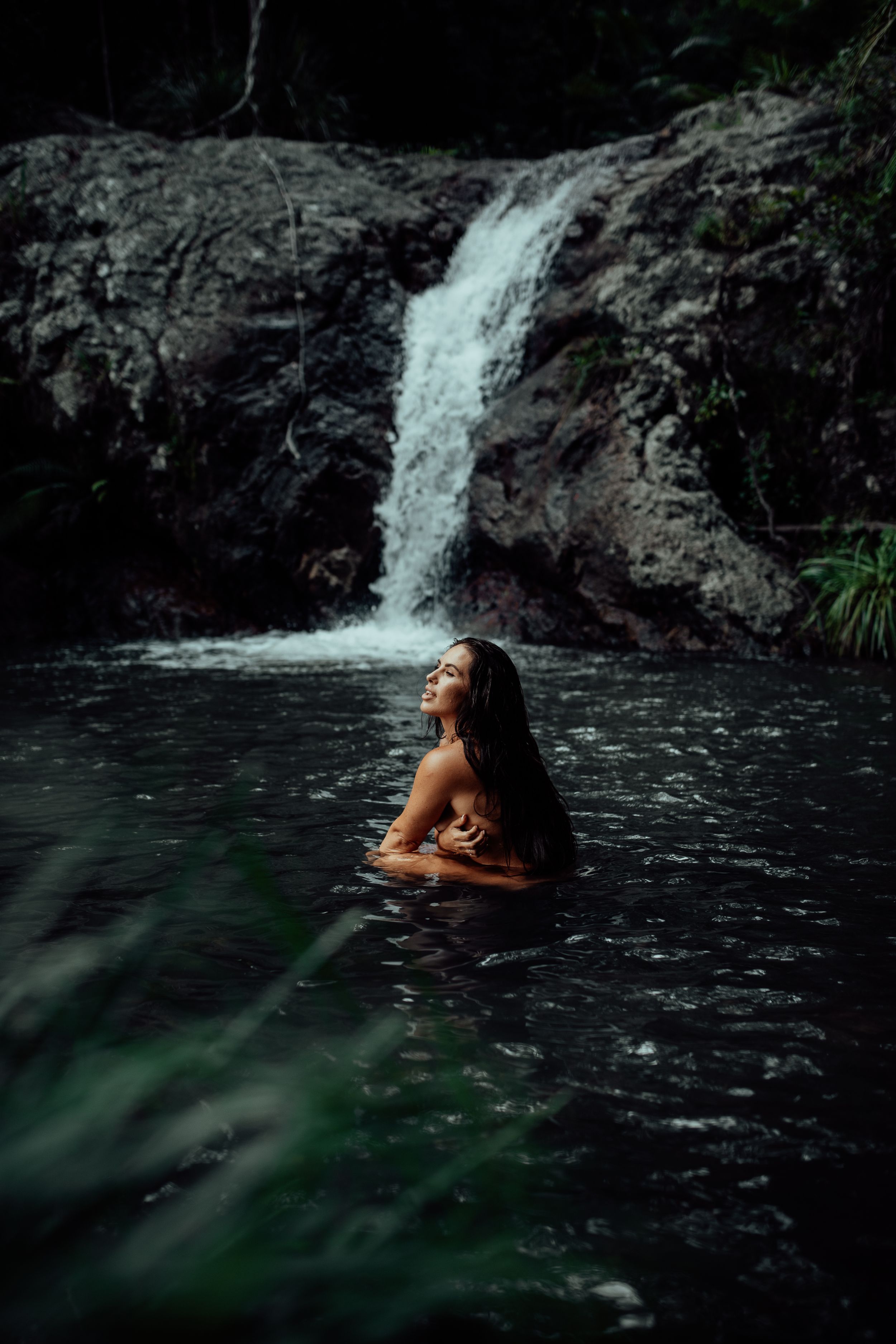 A woman bathes in a natural pool beneath a cascading waterfall in a moody forest setting.