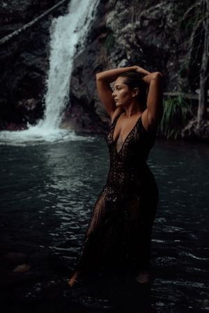 A person in a black lace dress poses dramatically in front of a forest waterfall.