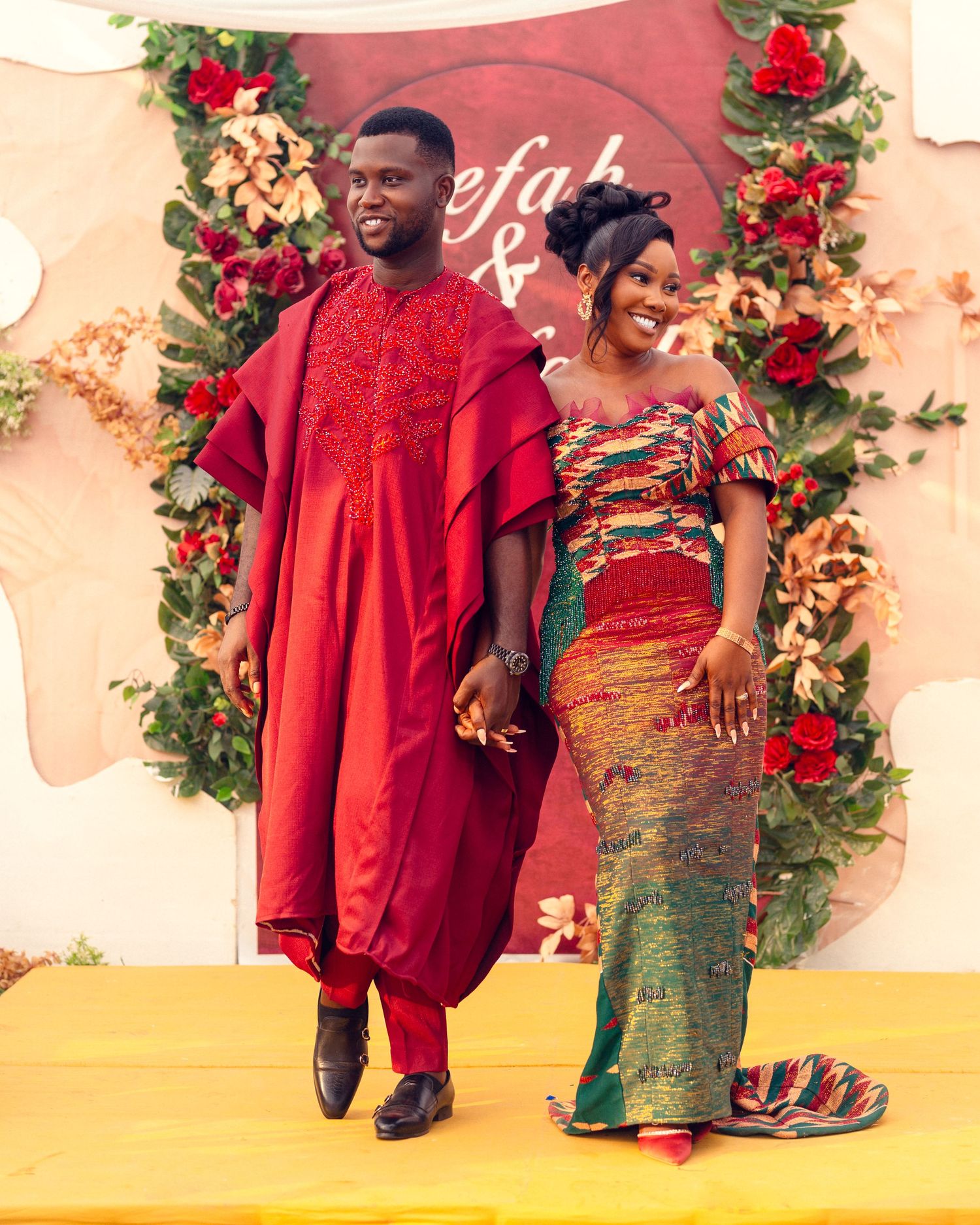 Couple in coordinated African attire poses on yellow stage with floral wall backdrop.