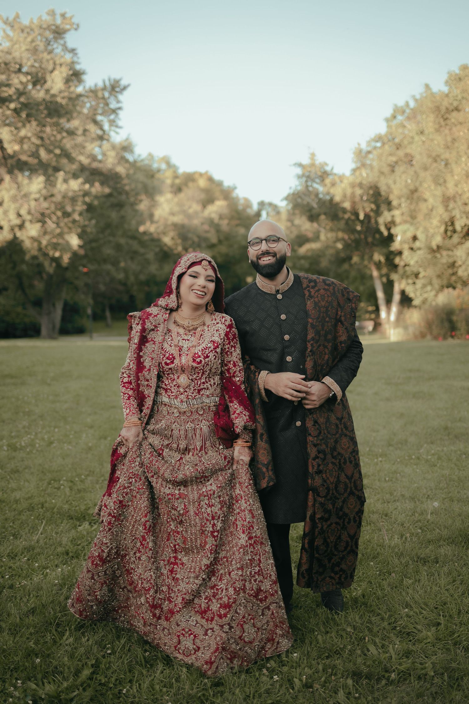 Couple in traditional South Asian wedding attire pose together in a sunlit garden setting.