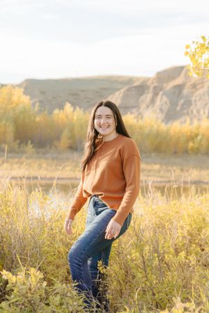 Person wearing terracotta sweater and jeans stands in tall grass with cliffs and autumn foliage behind.