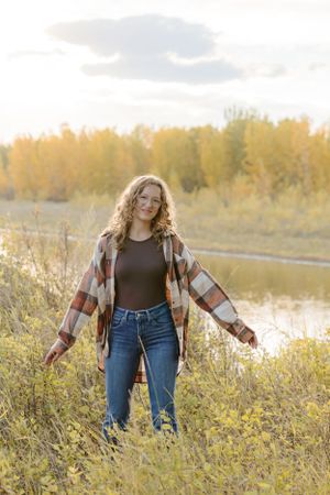 Person wearing plaid shirt and jeans poses in wild grass near water during sunset.