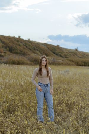 Person in beige top and light wash jeans stands in a golden field with hills in background.