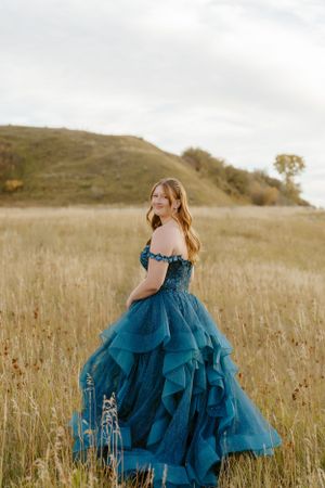 Figure wearing dramatic teal ballgown poses in autumn field against rolling hills.