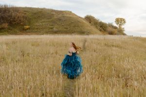A flowing blue ballgown dress stands out against golden prairie grass and rolling hills at sunset.