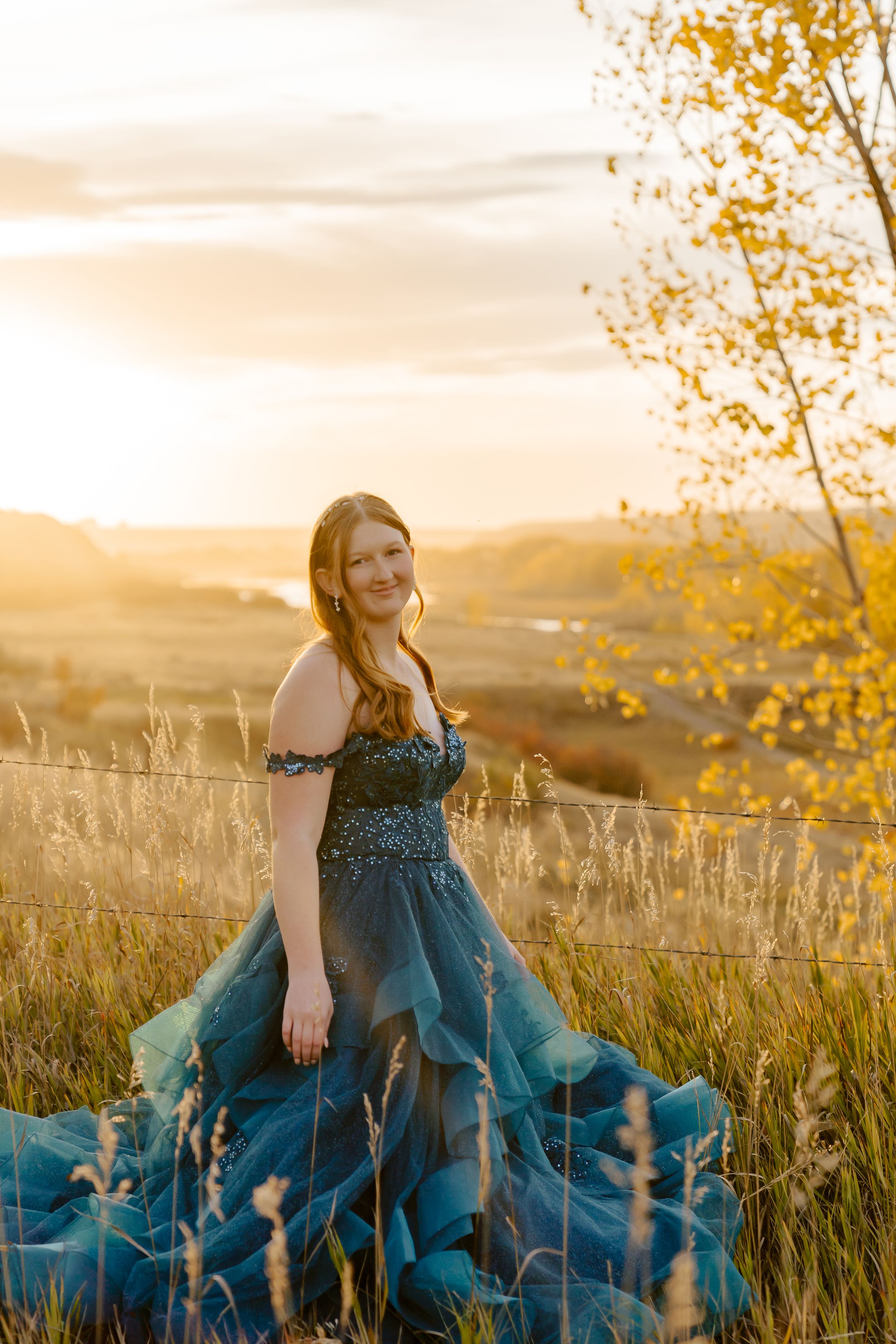 A blue formal gown catches the golden sunset light in a prairie field with autumn foliage.