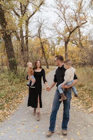 A family of four walks together on a leaf-covered path through a golden autumn forest.