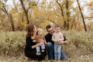 A family sits together on the ground surrounded by fall foliage and tall trees.