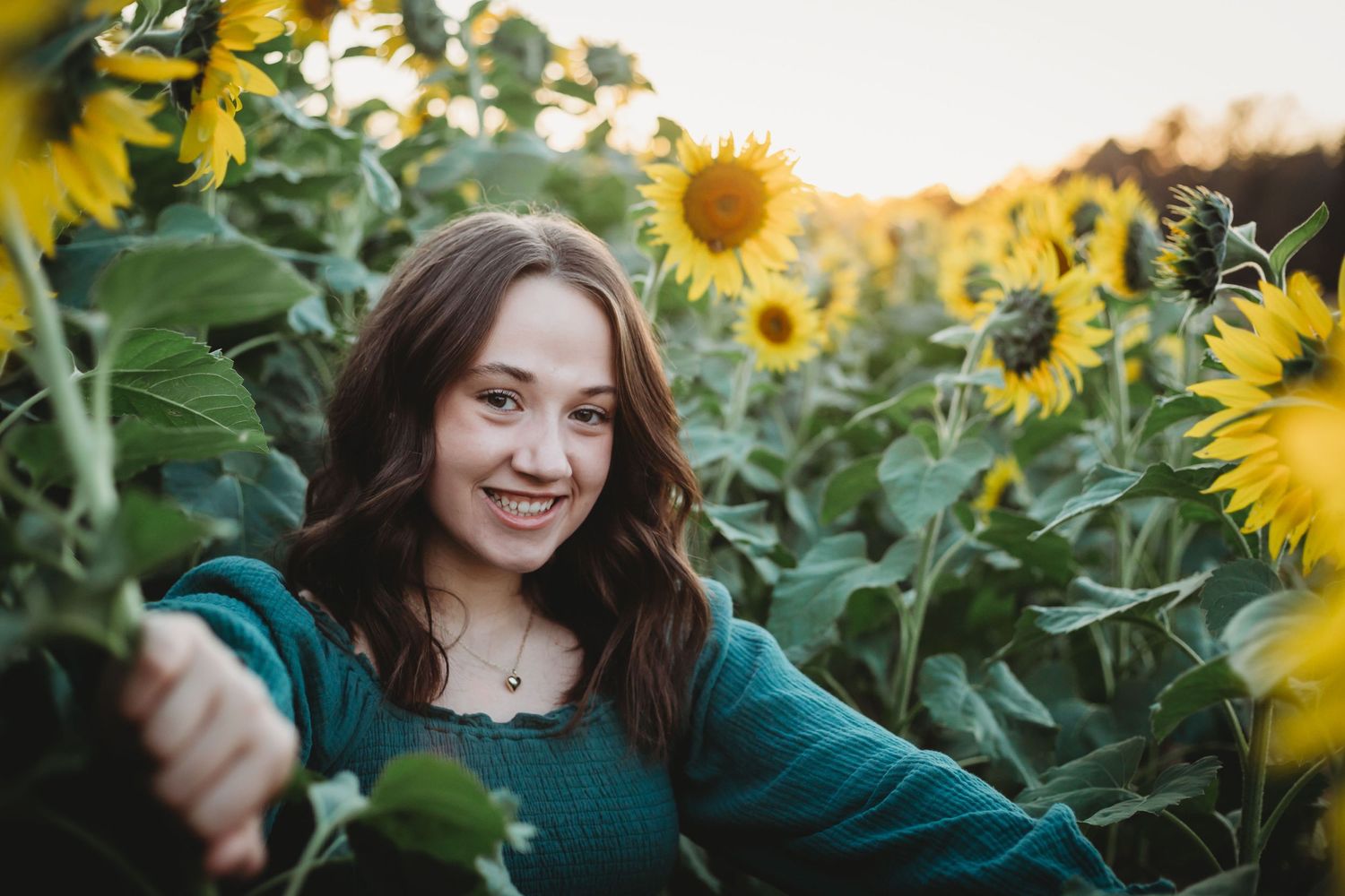 Someone in a green top smiles while standing in a vibrant field of tall blooming sunflowers during golden hour.