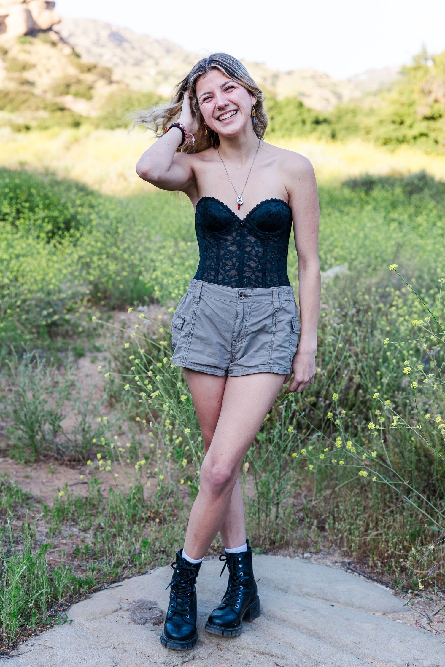 Someone poses outdoors wearing a black corset top, khaki shorts and combat boots in a natural setting.