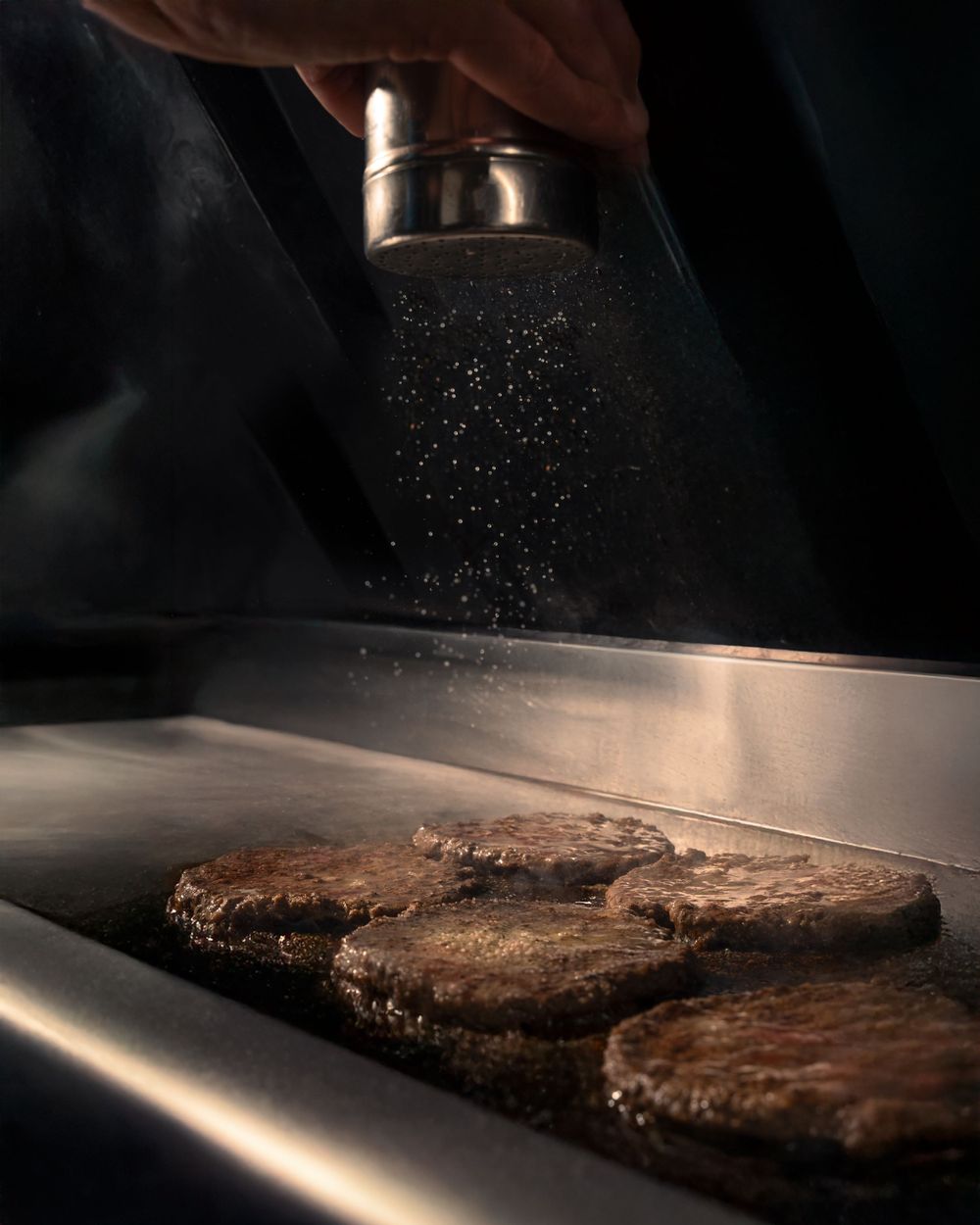 Seasoning being sprinkled on burger patties cooking on a dark grill surface. Mtlshutter photography
