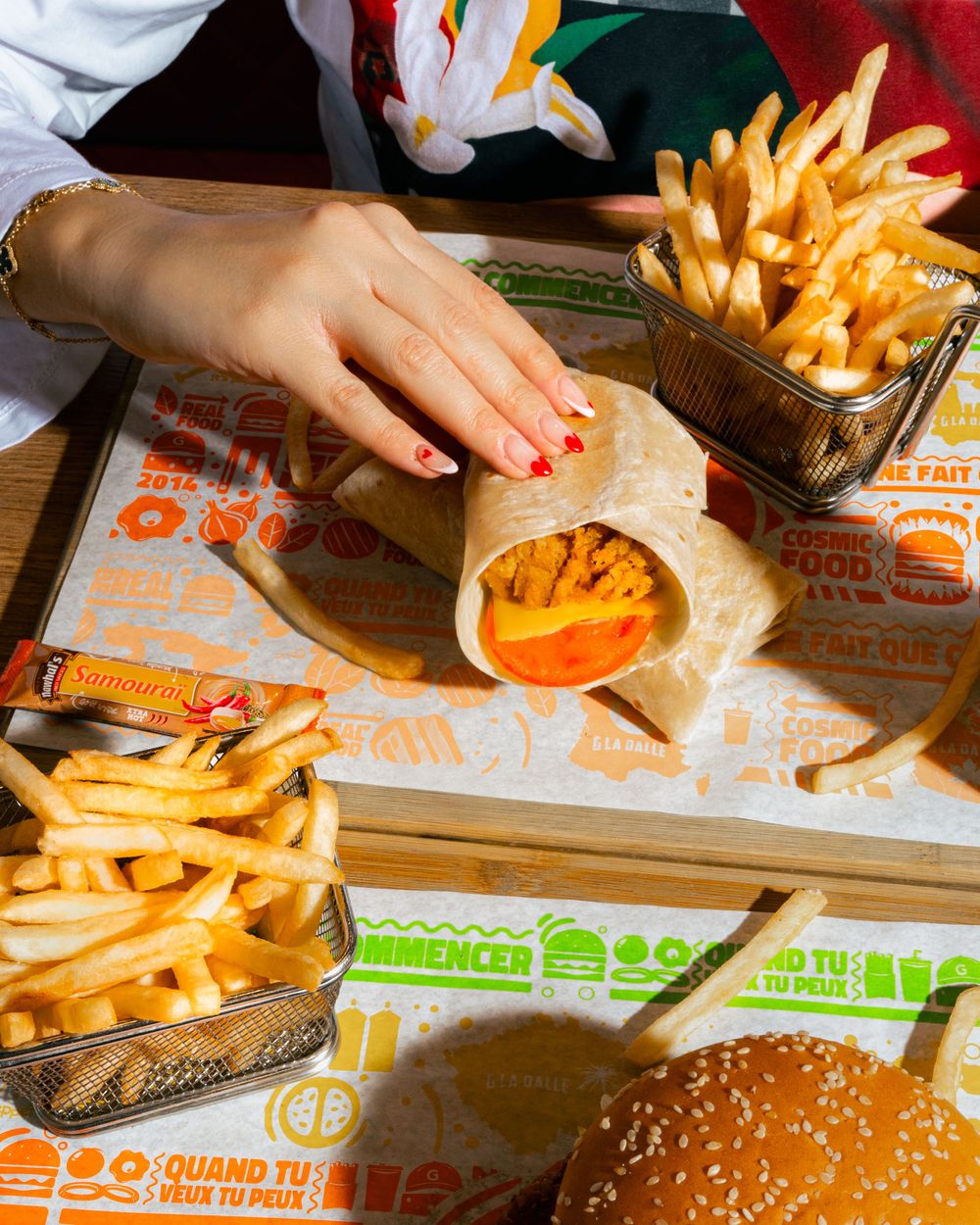 A hand reaches for a wrapped burger and fries on orange branded paper with colorful text. Mtlshutter photography