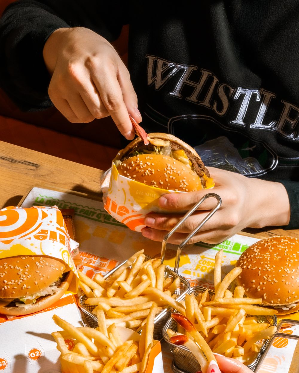 Hands unwrapping a cheeseburger with multiple burgers and fries on wooden table. Mtlshutter photography
