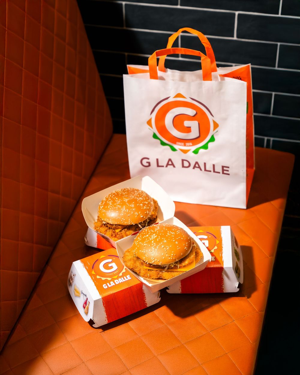 Orange and black branded Grille burger packaging with a hamburger displayed on orange surface by dark tile wall.