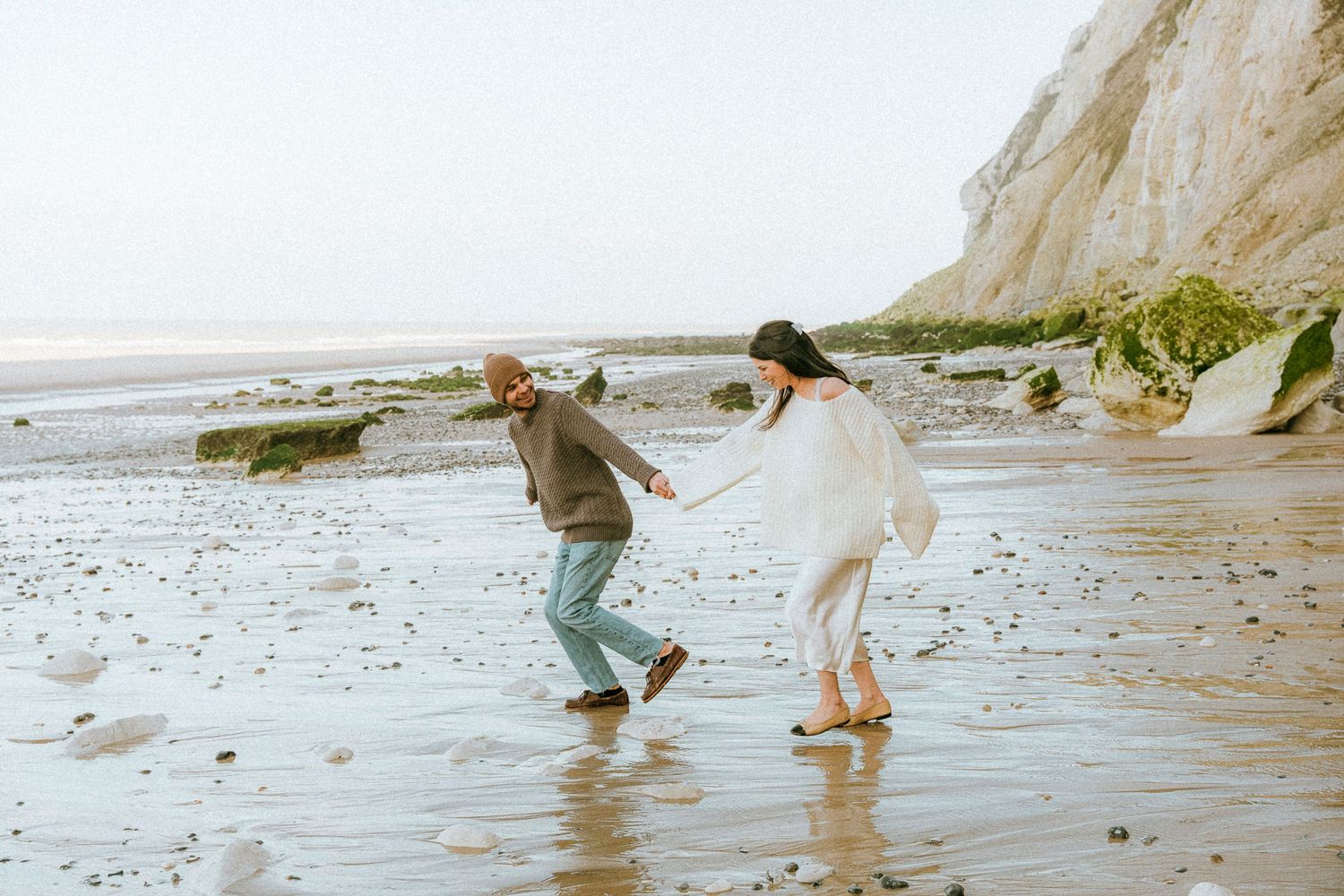 Two people dance and splash joyfully along a scenic beach with white cliffs in the background.