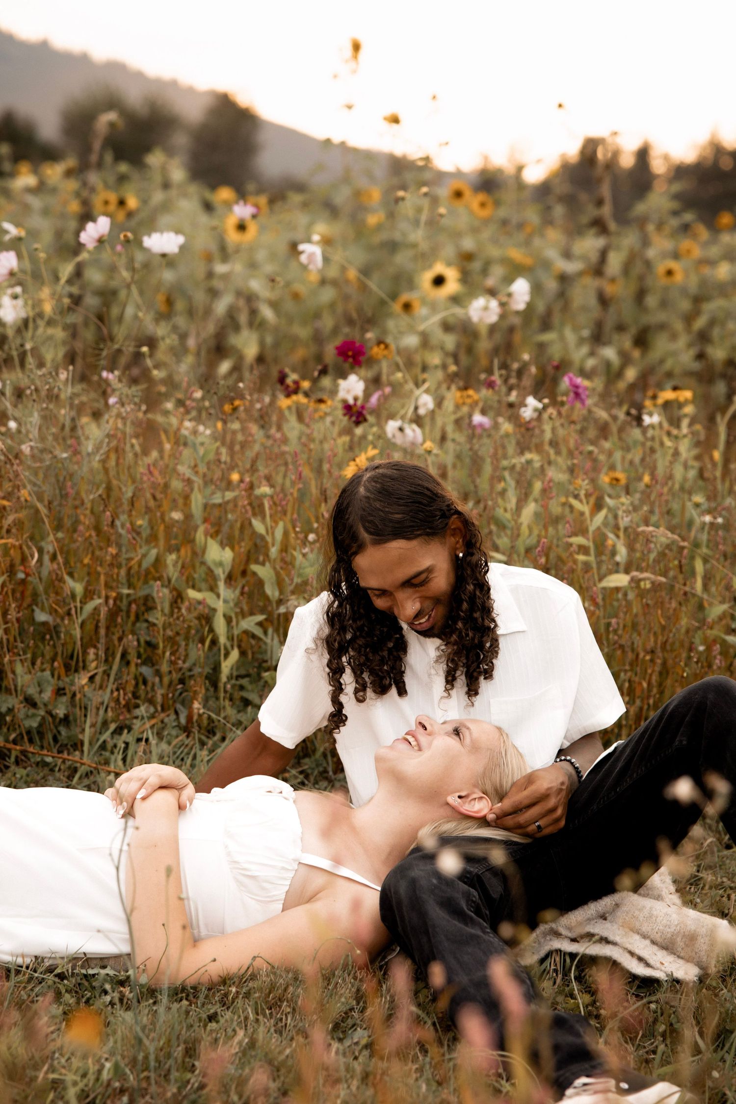 Couple relaxing in a wildflower field at sunset, woman lying in mans lap smiling, sunflowers blooming in background.