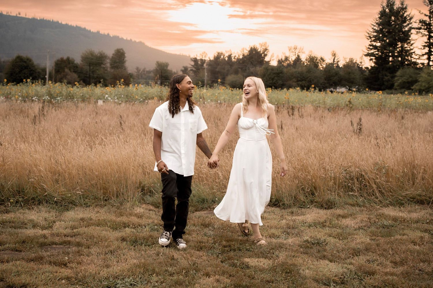 Couple holding hands walking through golden field at sunset, woman in white dress, man in white shirt and black pants.