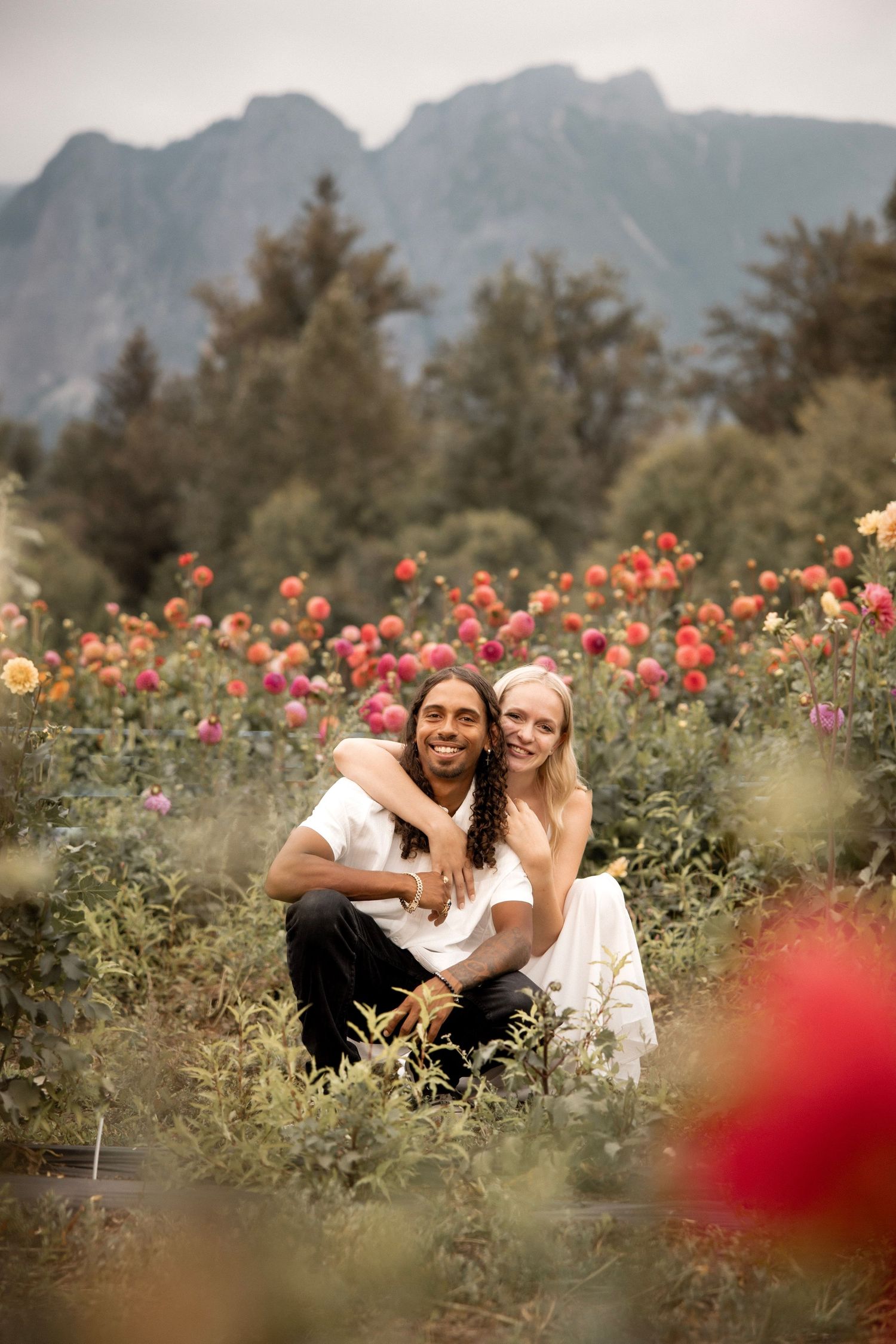 Couple sitting together in a vibrant dahlia flower field with mountains and trees in the background.