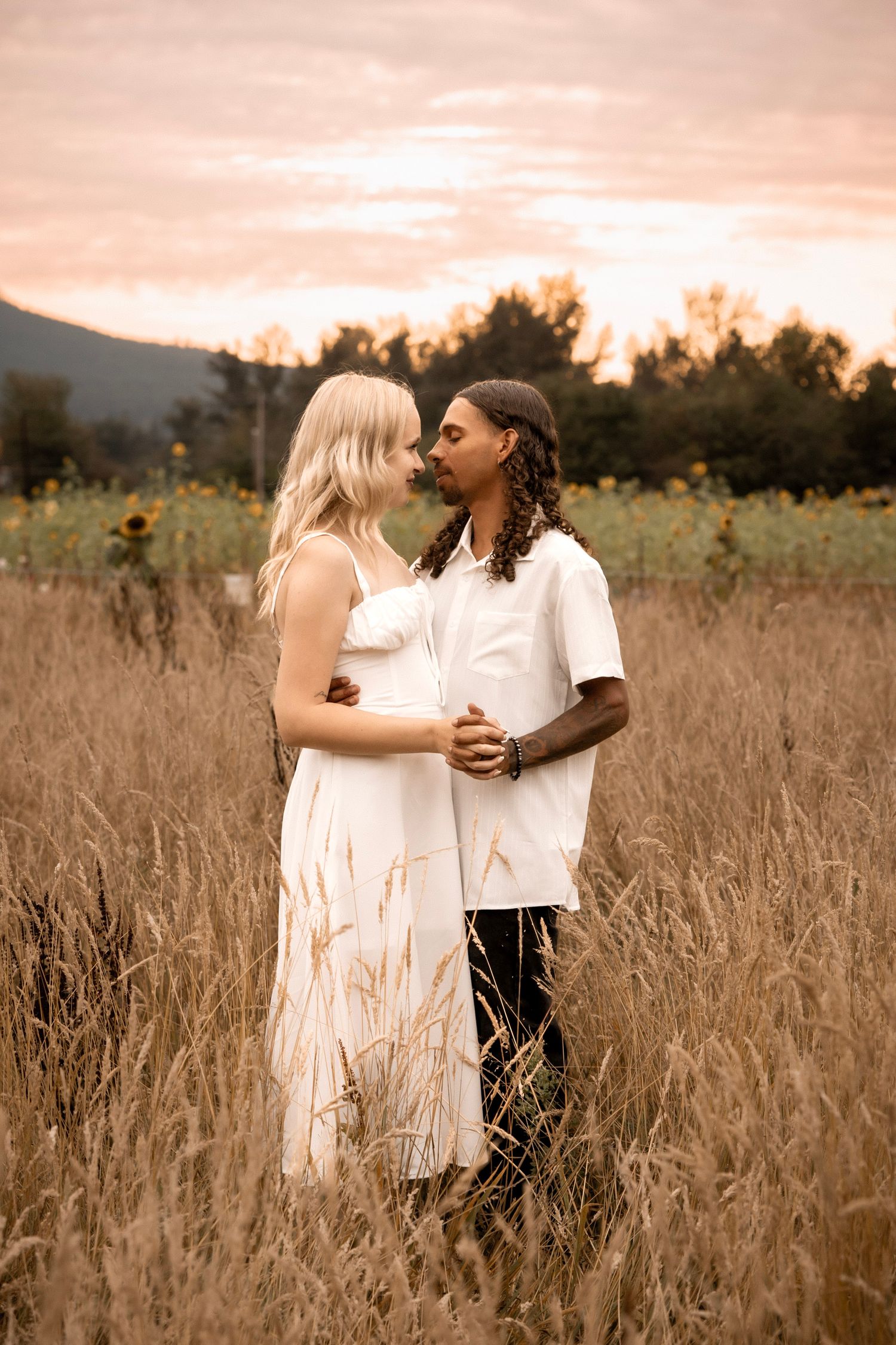 Couple embracing in golden field at sunset, woman in white dress, man in white shirt, sunflowers and mountains behind.