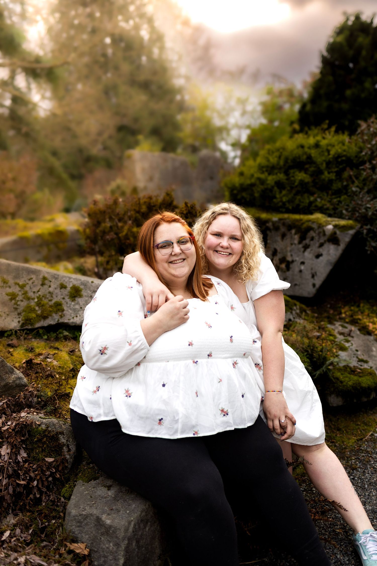 Two people in white tops sitting together on rocky terrain in a garden setting.