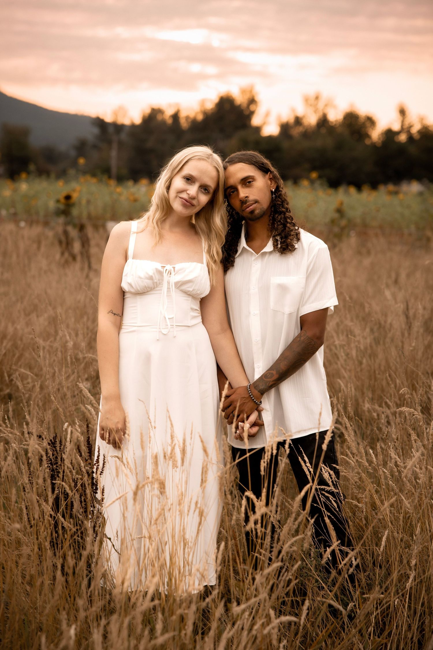 A couple holds hands in a golden field at sunset, woman in white dress, man in white shirt, trees in background.