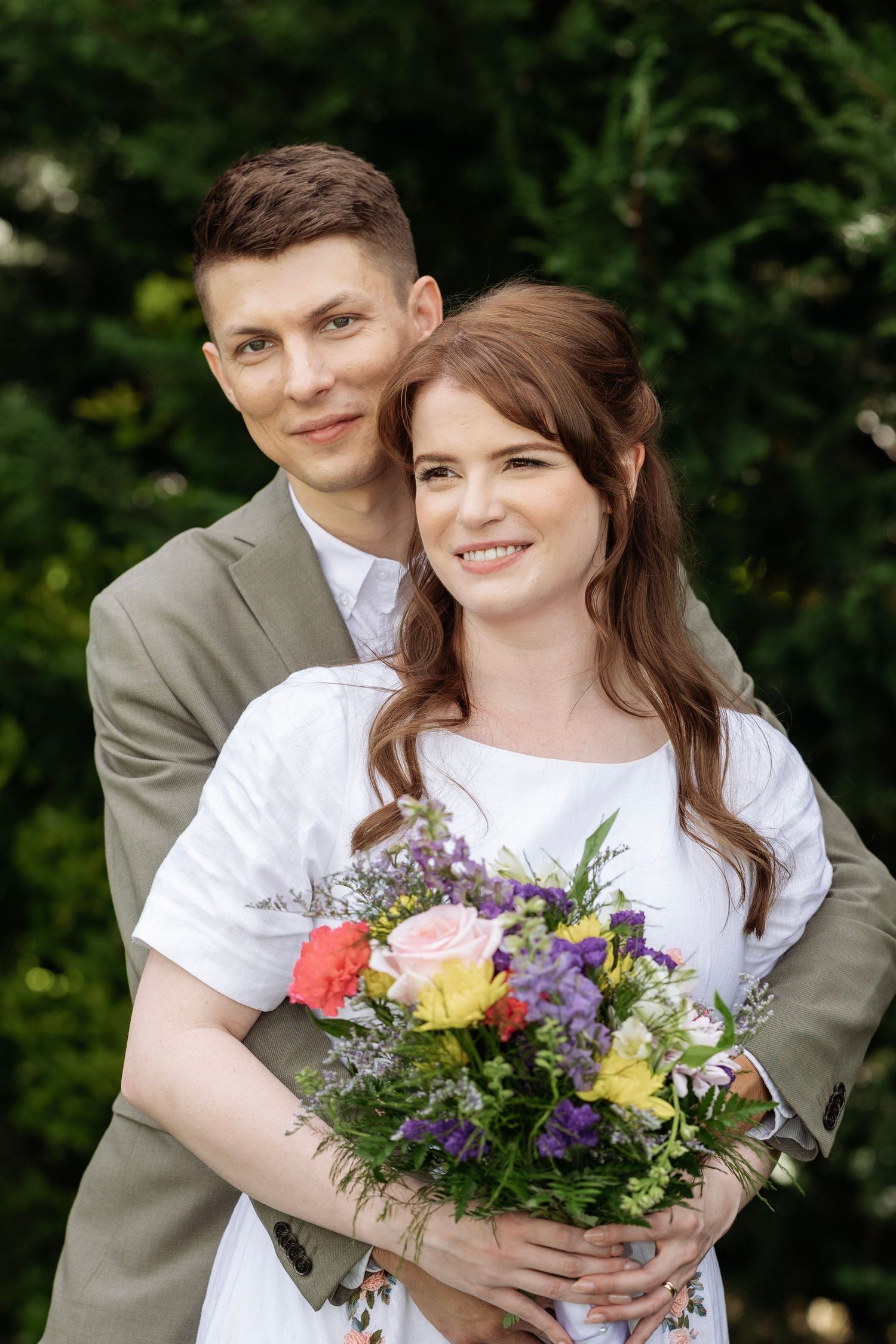 Happy couple posing outdoors, bride in white dress holding colorful bouquet, groom in gray suit embracing her.