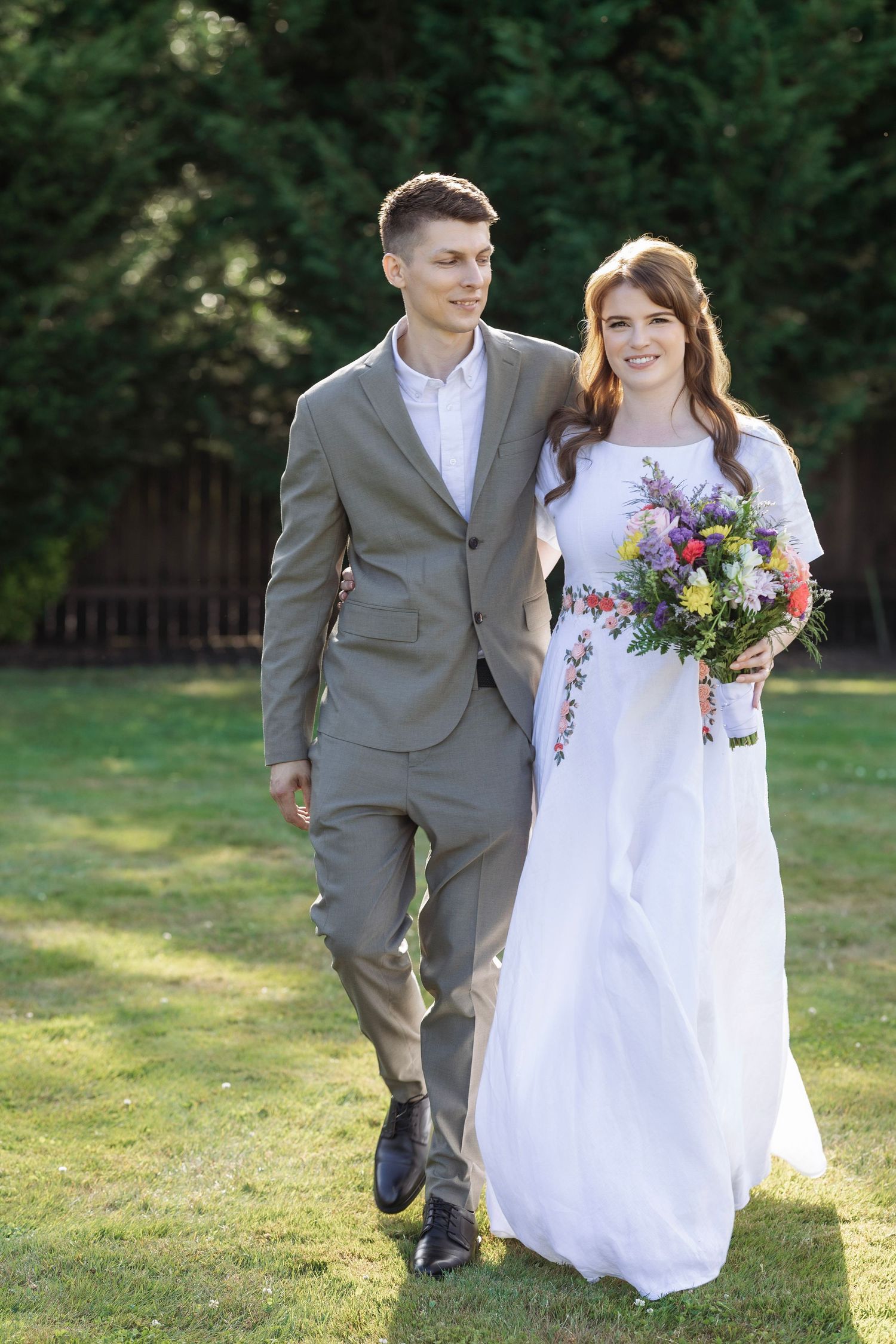 A smiling bride in a white gown holding a colorful bouquet walks with a groom in a gray suit on a sunny lawn.