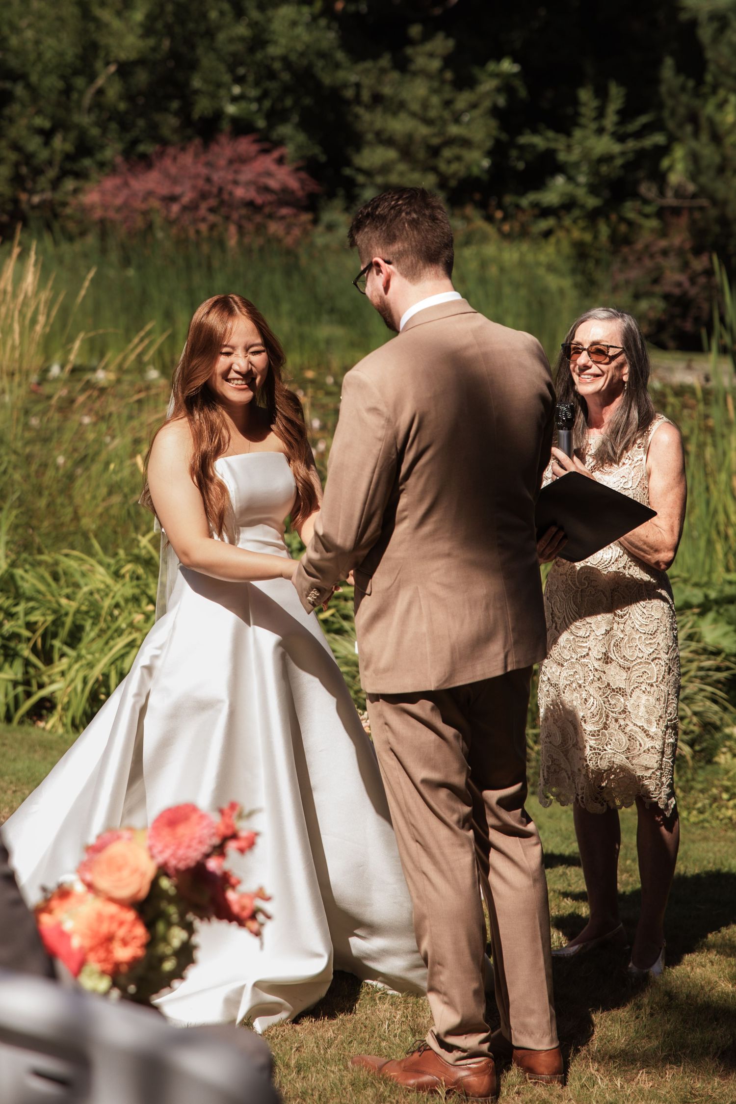 Bride smiling joyfully at groom during outdoor wedding ceremony with officiant holding book in garden setting.