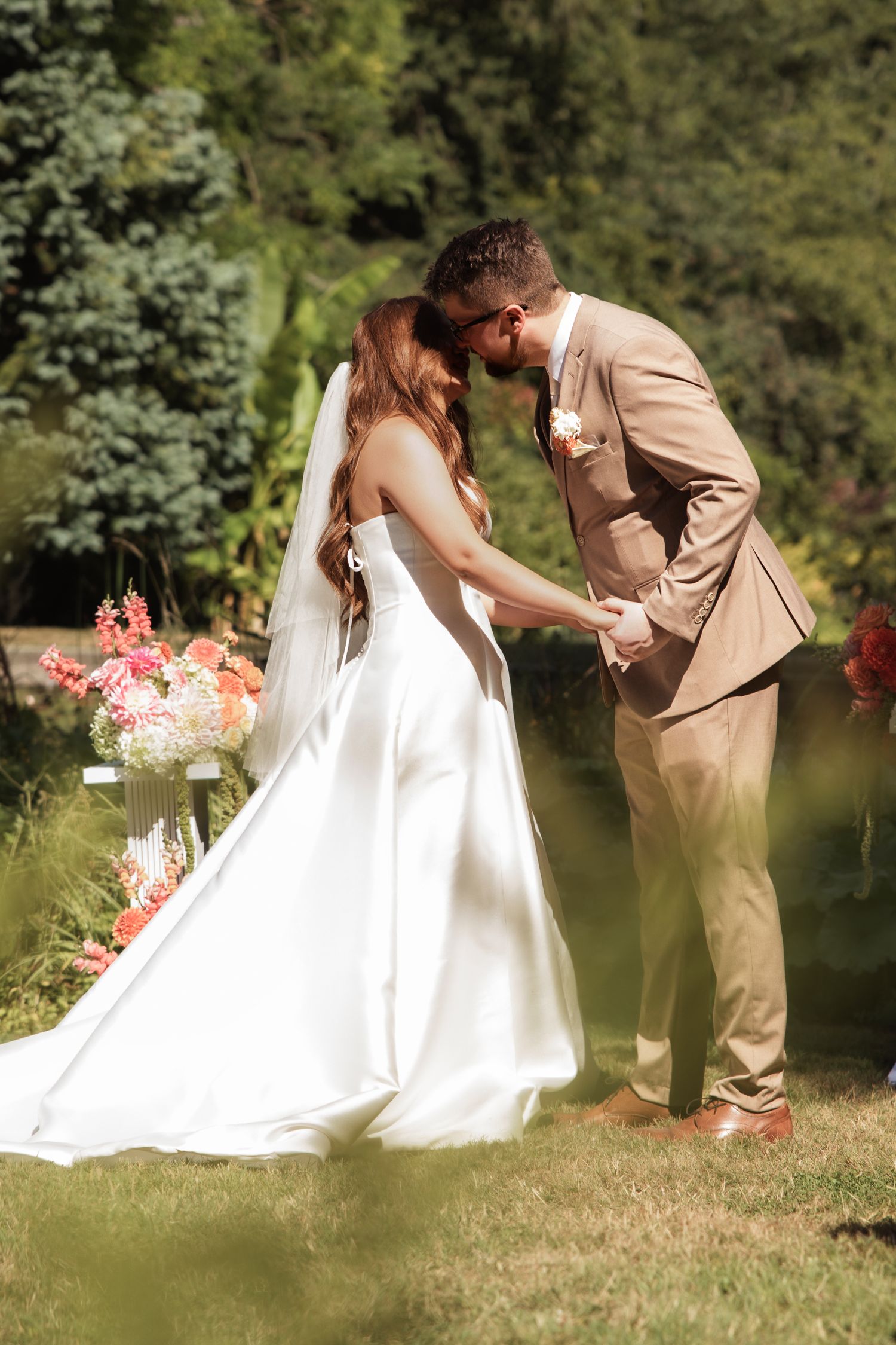 Bride and groom share a romantic kiss while holding hands during their outdoor garden wedding ceremony.