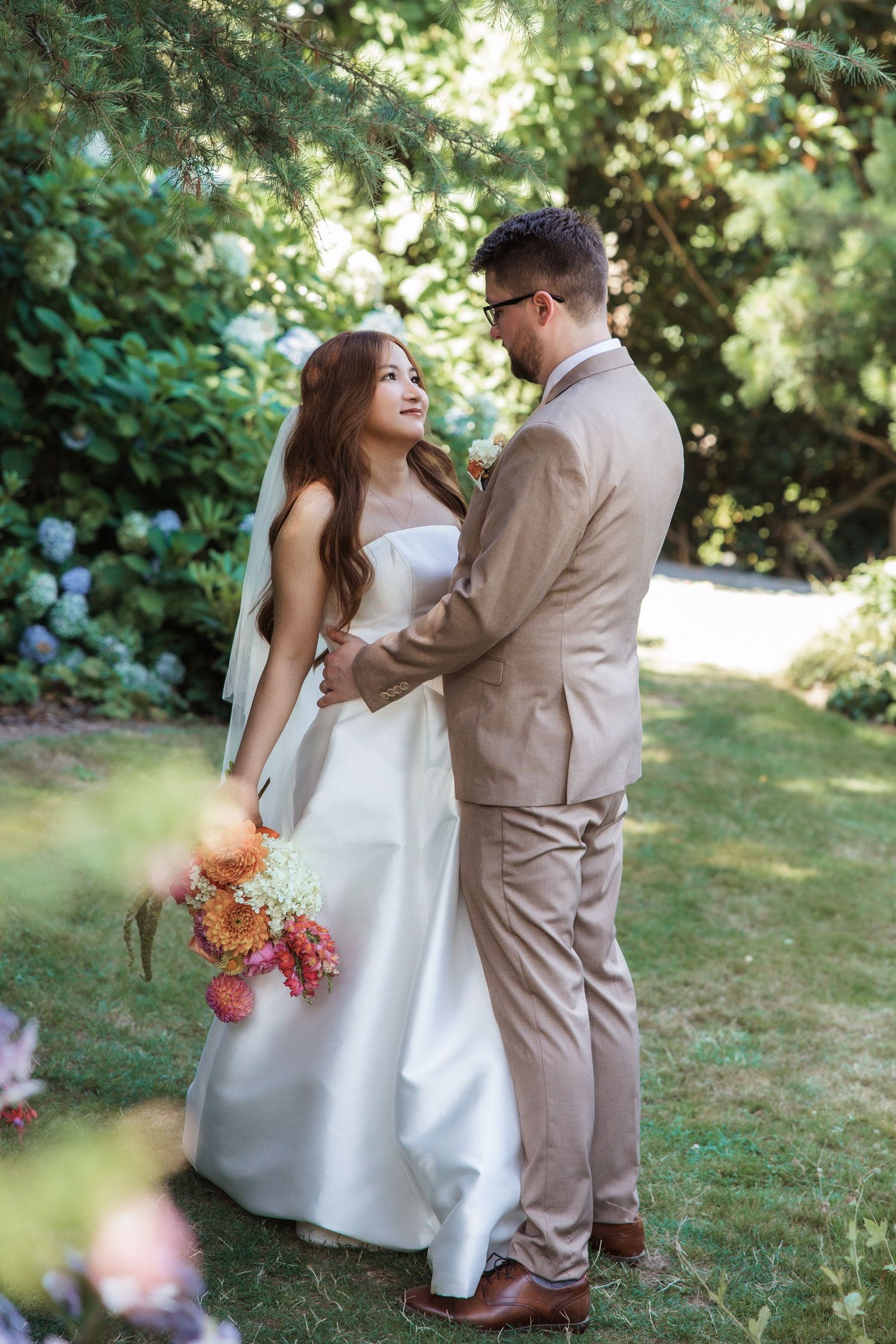 Smiling bride in white gown holds colorful bouquet while standing with groom in tan suit in garden.