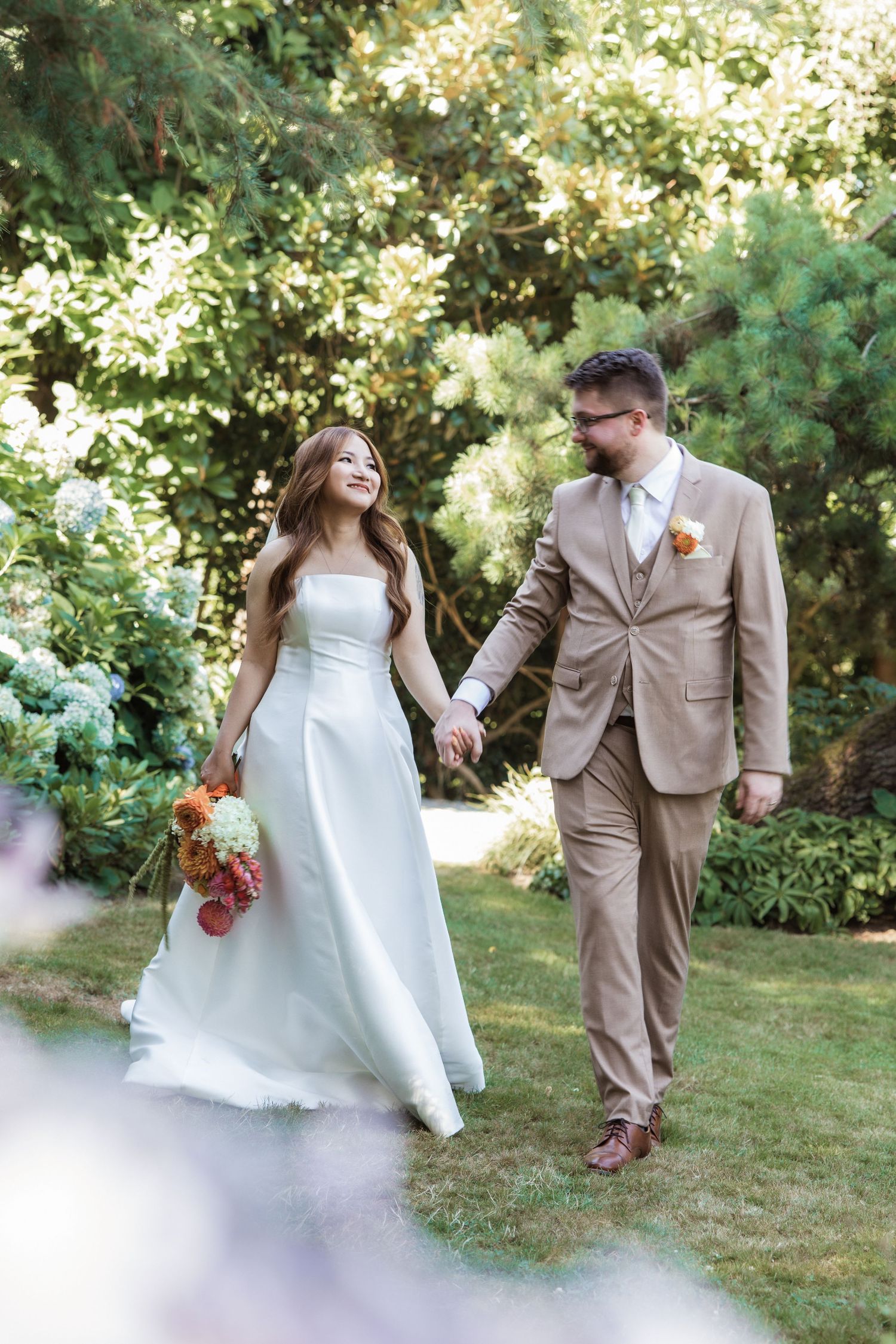 Bride and groom walking hand in hand through lush garden, bride holding colorful bouquet in elegant white gown.