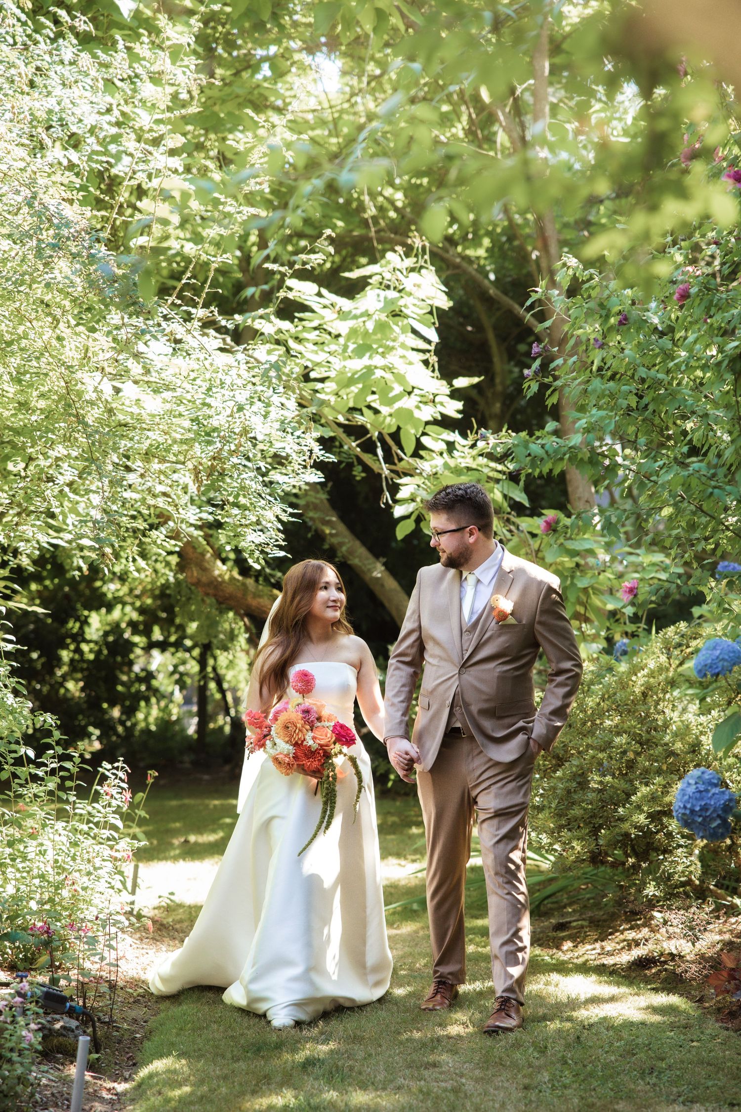 A bride and groom walk hand-in-hand through a lush garden, she holds a vibrant bouquet in her white gown.