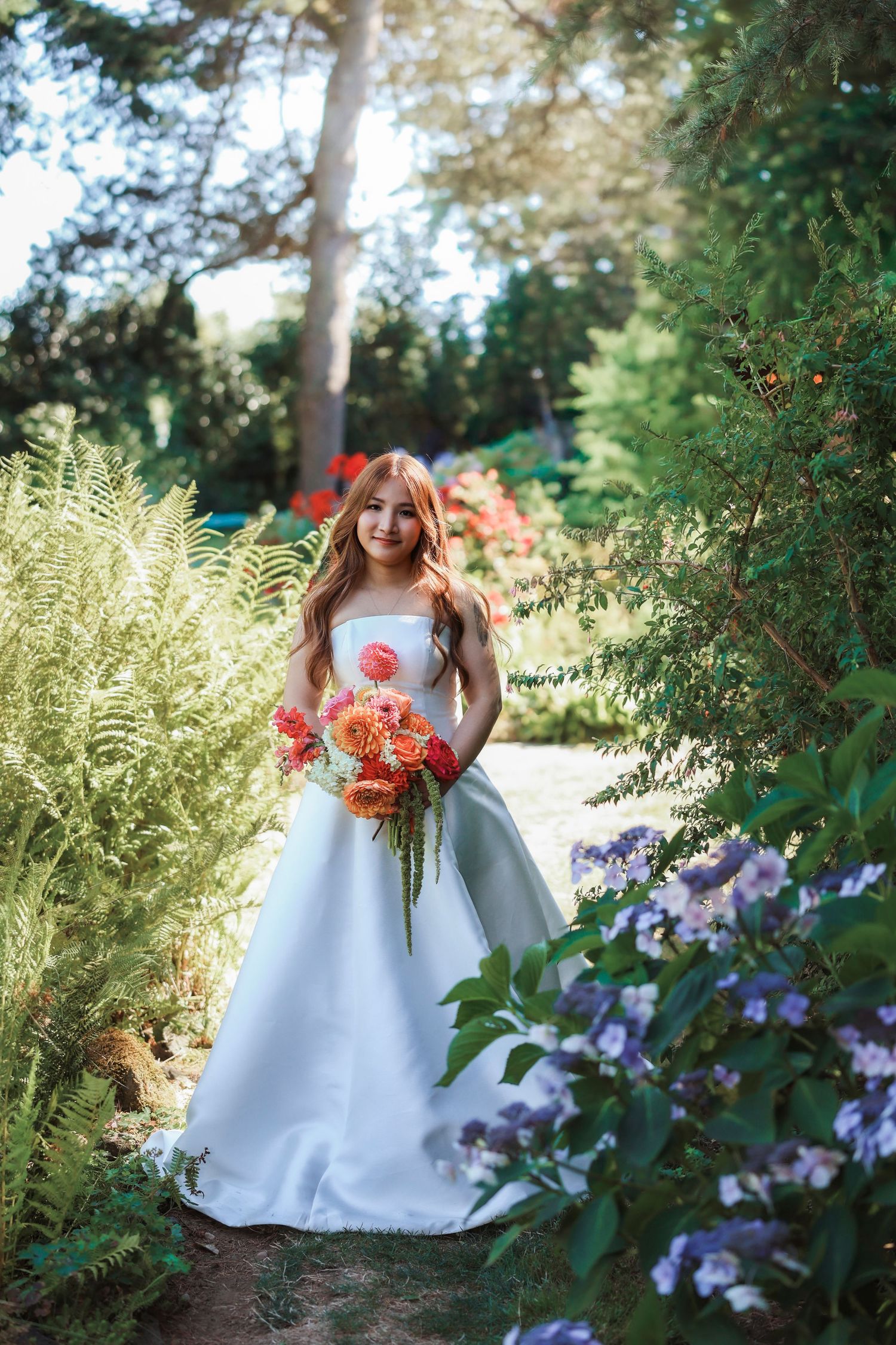 Smiling bride in white gown holding colorful bouquet surrounded by lush garden greenery and blooming flowers.
