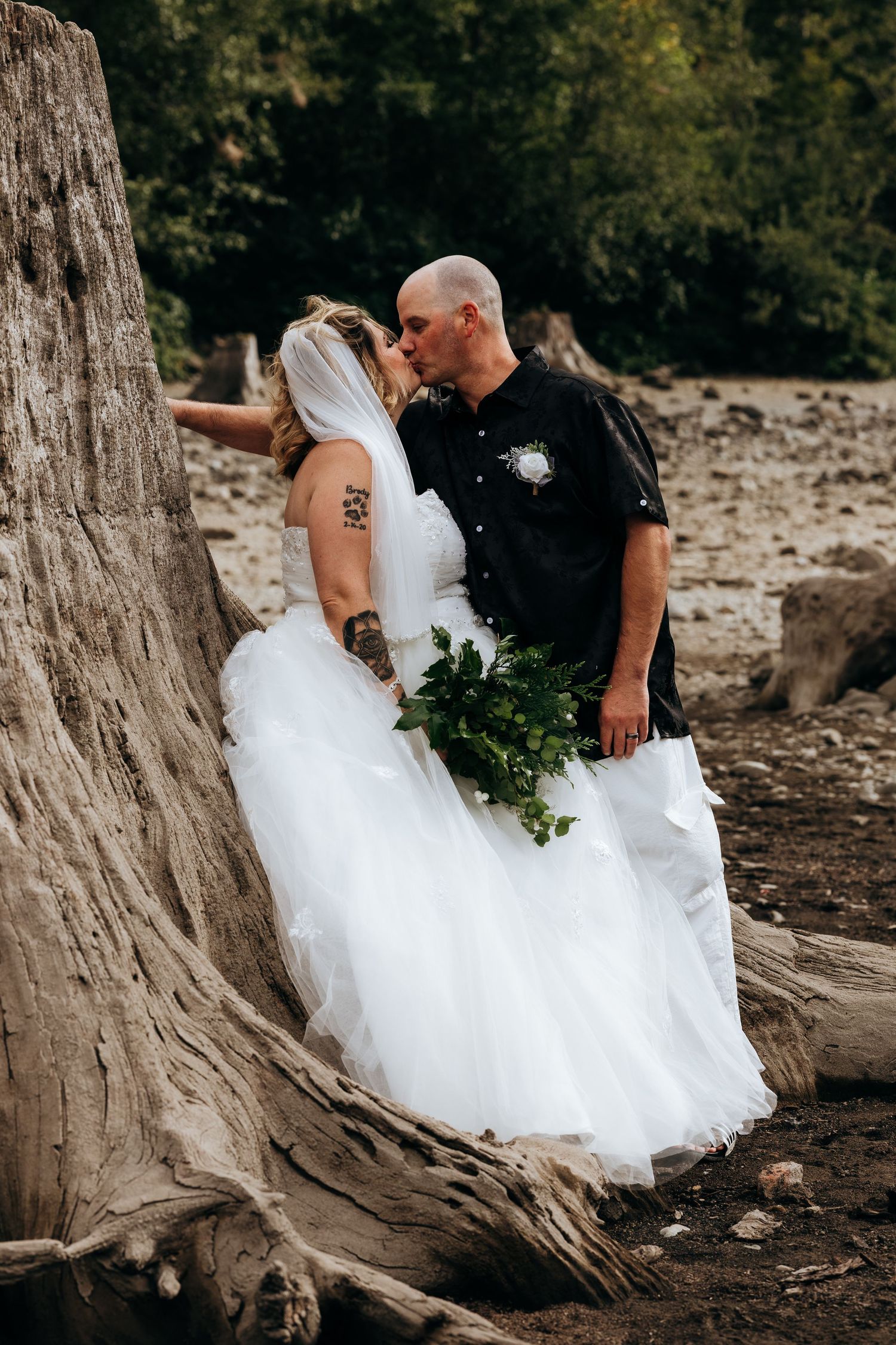 A couple shares an intimate moment on a fallen log by the water's edge during their wedding photoshoot.