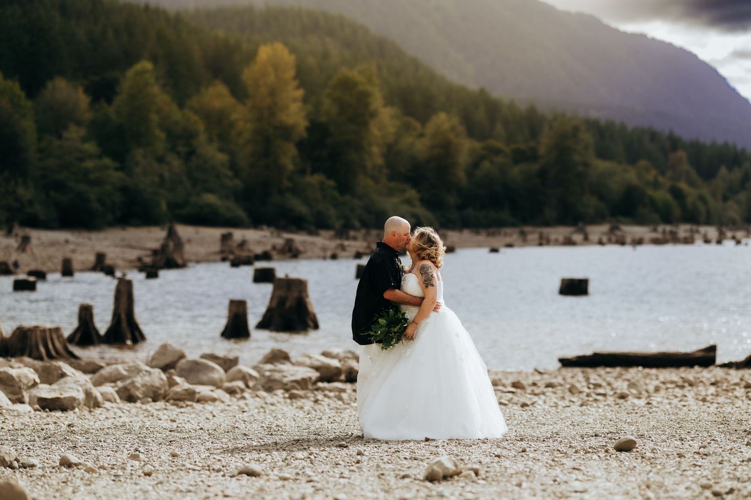 Couple sharing an intimate moment on a scenic rocky beach with mountains and lake in the background.