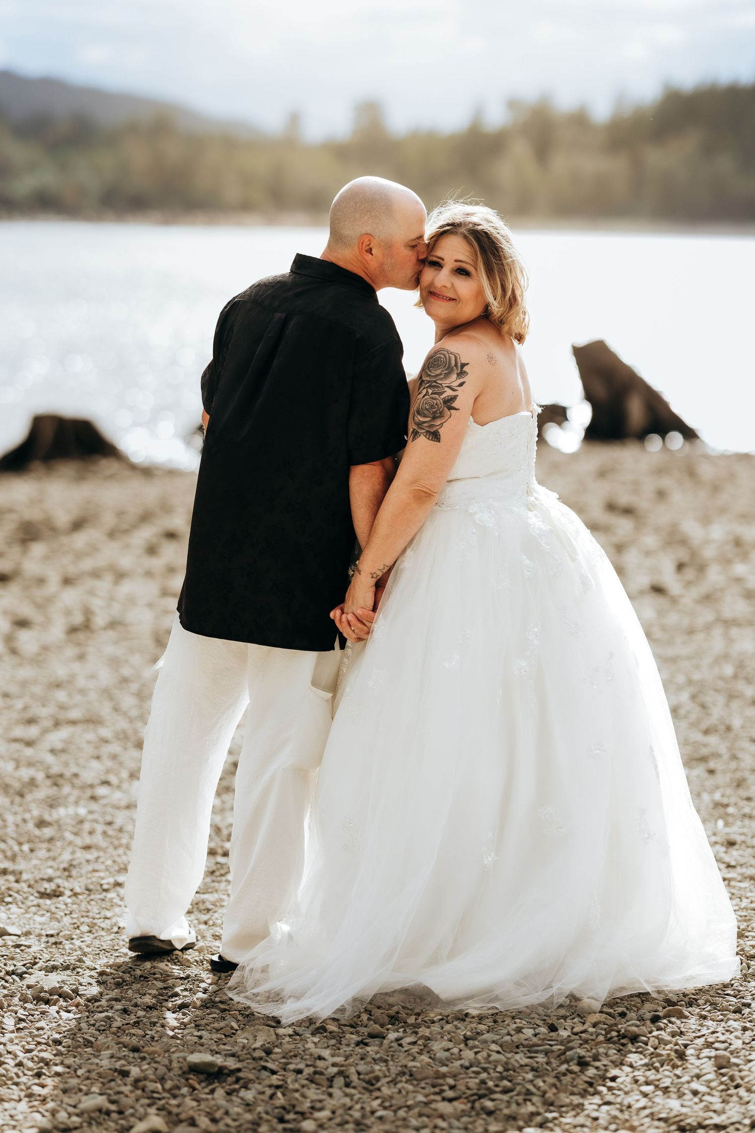 A couple in wedding attire poses together on a rocky beach shore with water in the background.